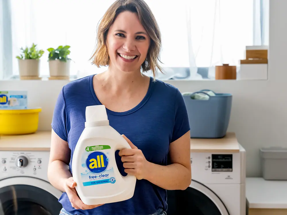 Woman smiling in front of washing machine and dryer holding a bottle of laundry detergent