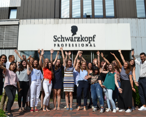 Ett mångsidigt Henkel-team som står och hejar framför Schwarzkopfs professionella byggnad och höjer sina armar