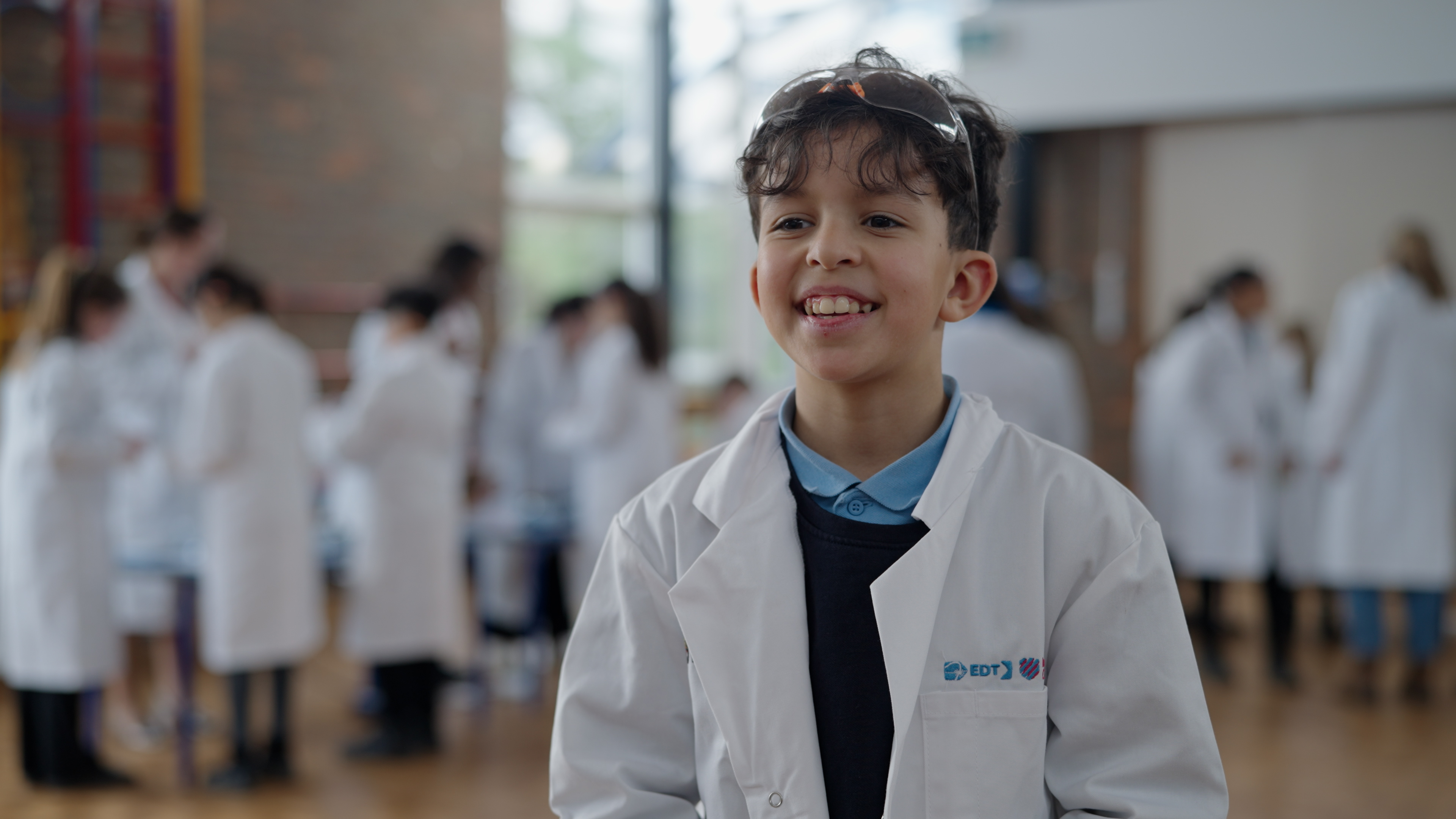 A young boy wearing a white lab coat stands and smiles brightly. He appears excited and engaged.