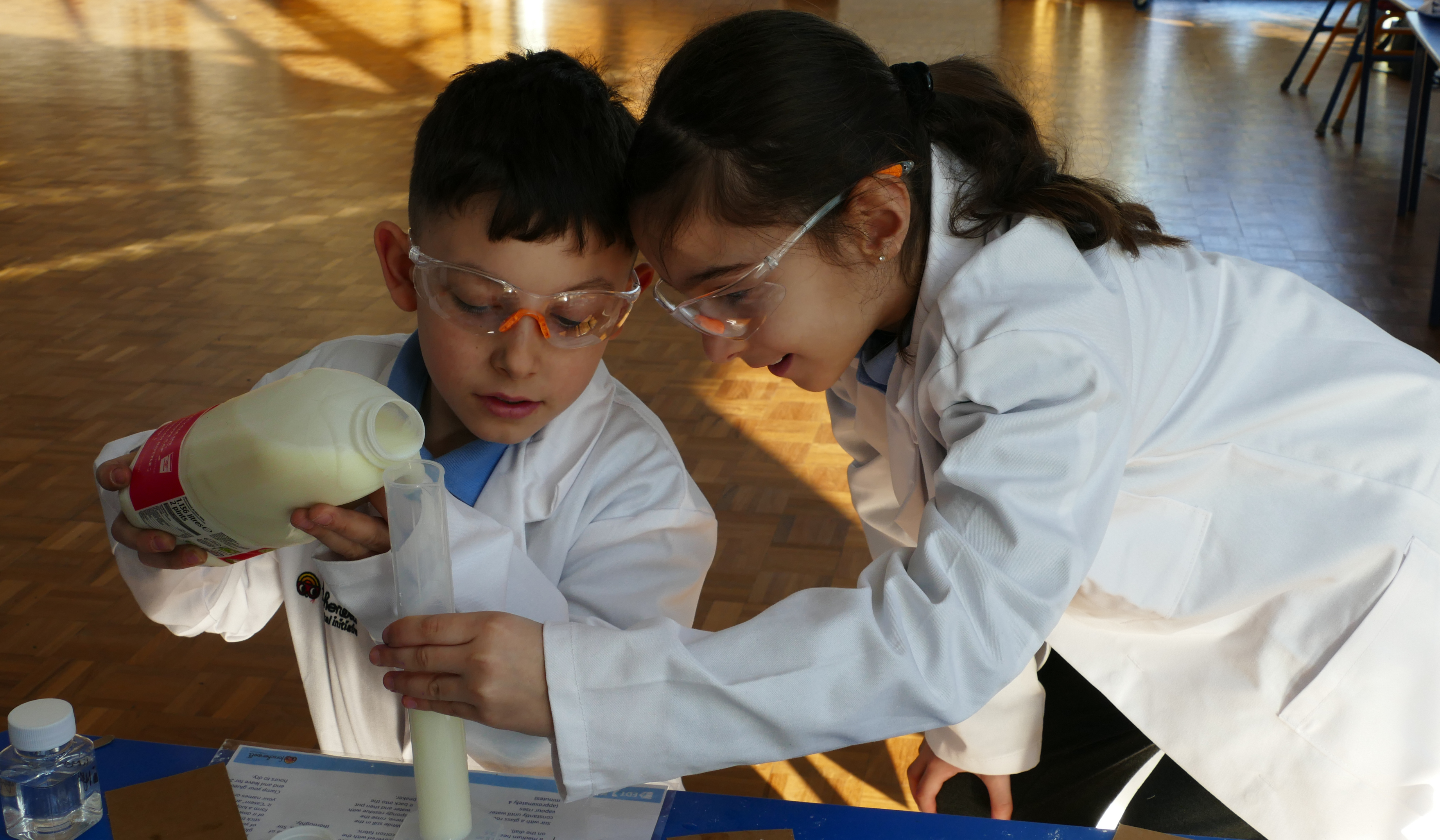 A young boy and girl wearing white lab coats and safety goggles conduct a science experiment.