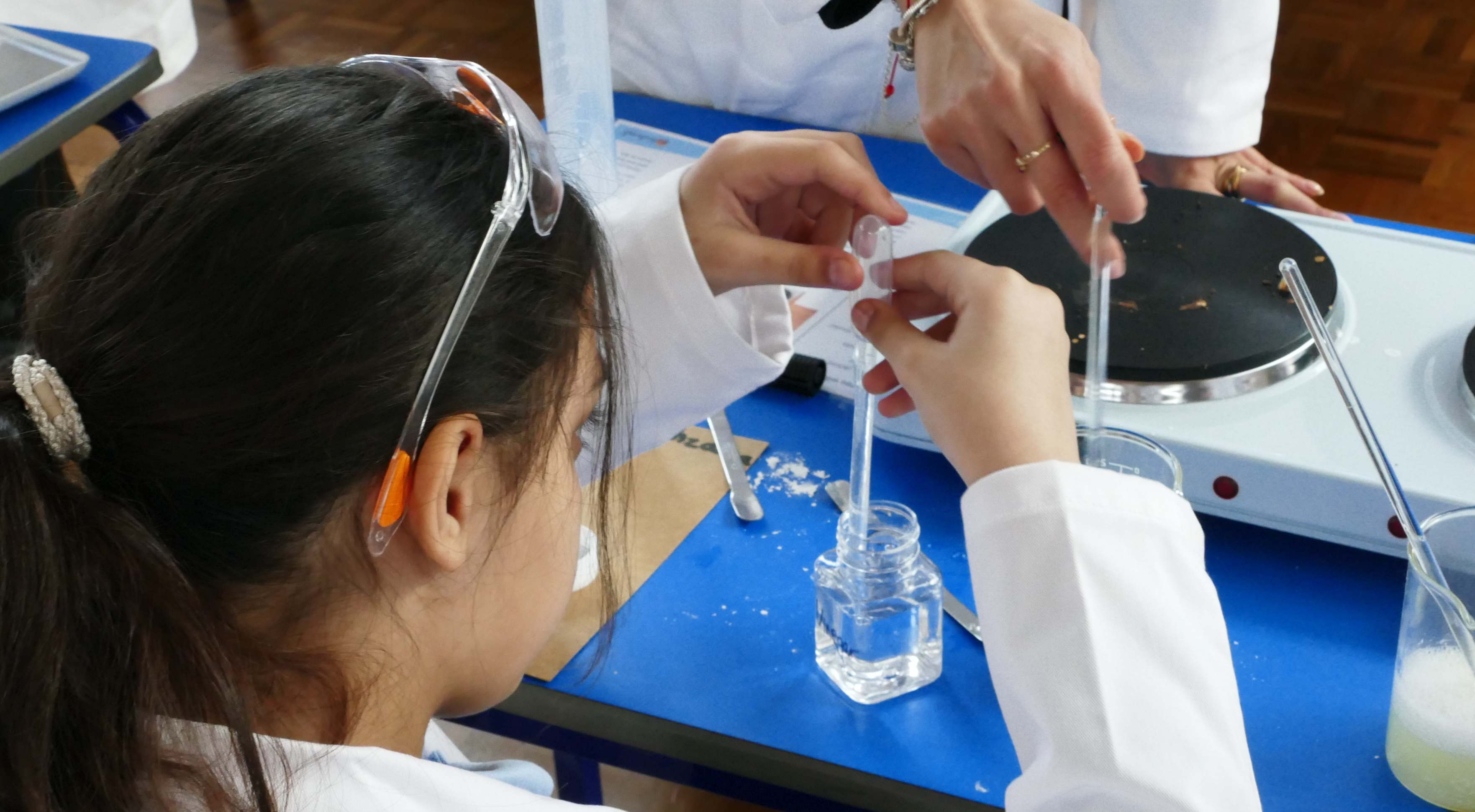 Two young boys wearing white lab coats and safety goggles conduct a science experiment.