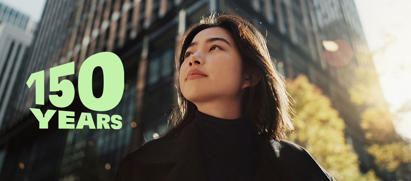  Green text reading ‘150 YEARS’ over a modern cityscape with tall glass buildings and autumn trees, symbolizing the 150th anniversary celebration.