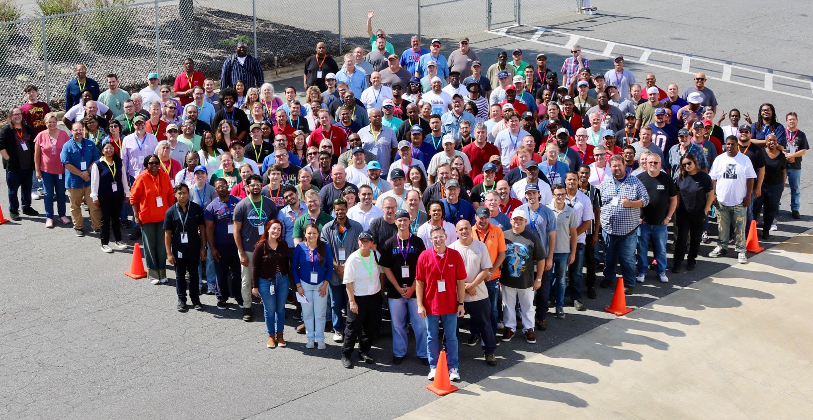 Group photo of employees arranged in a triangular formation between orange traffic cones in a parking lot.
