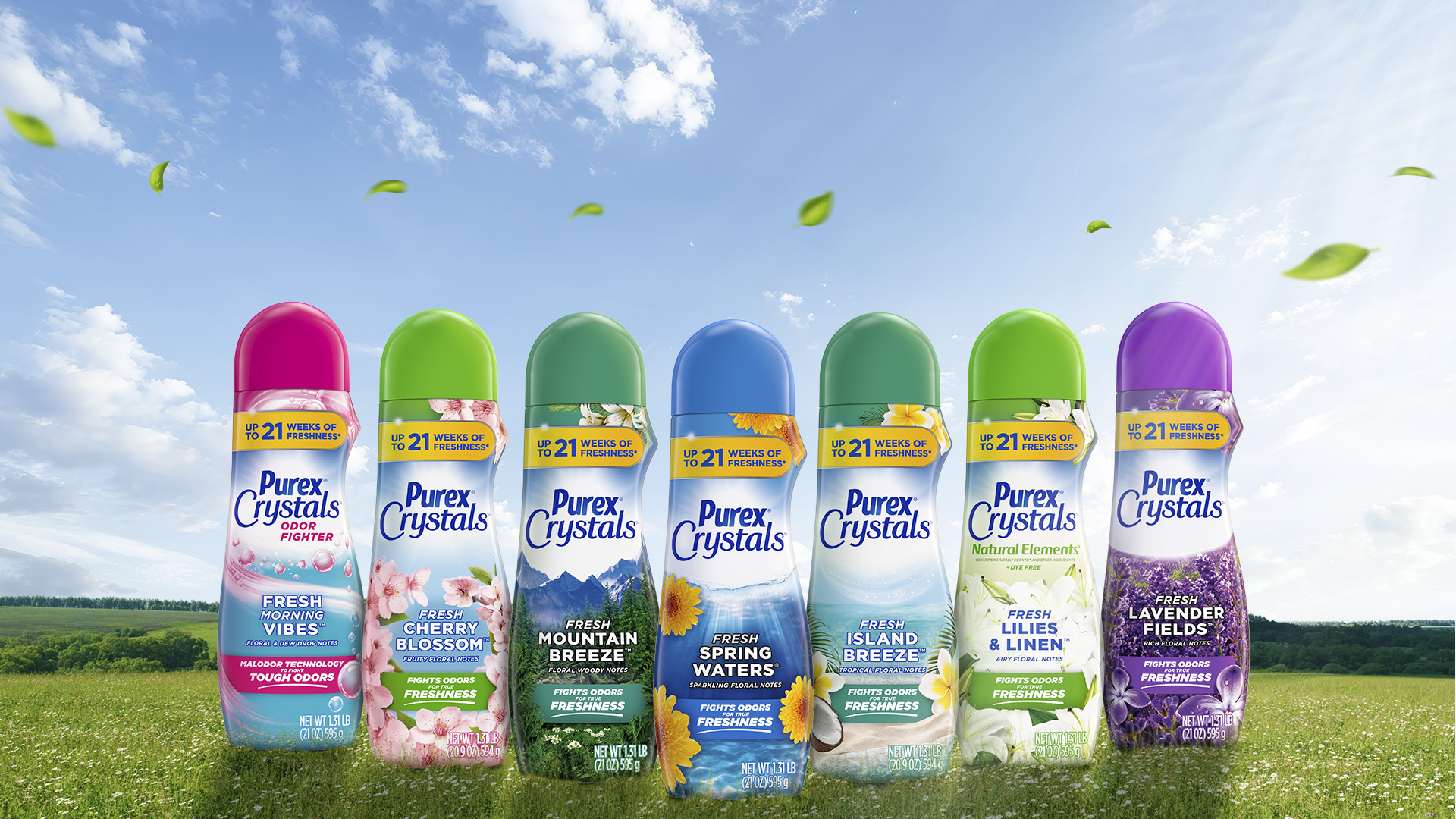 Seven Purex Crystals in-wash scent booster bottles standing on grass under a bright blue sky, showing different varieties including Fresh Vibes, Cherry Blossom, Mountain Breeze, Spring Waters, Island Breeze, Lilies & Linen, and Lavender Fields.