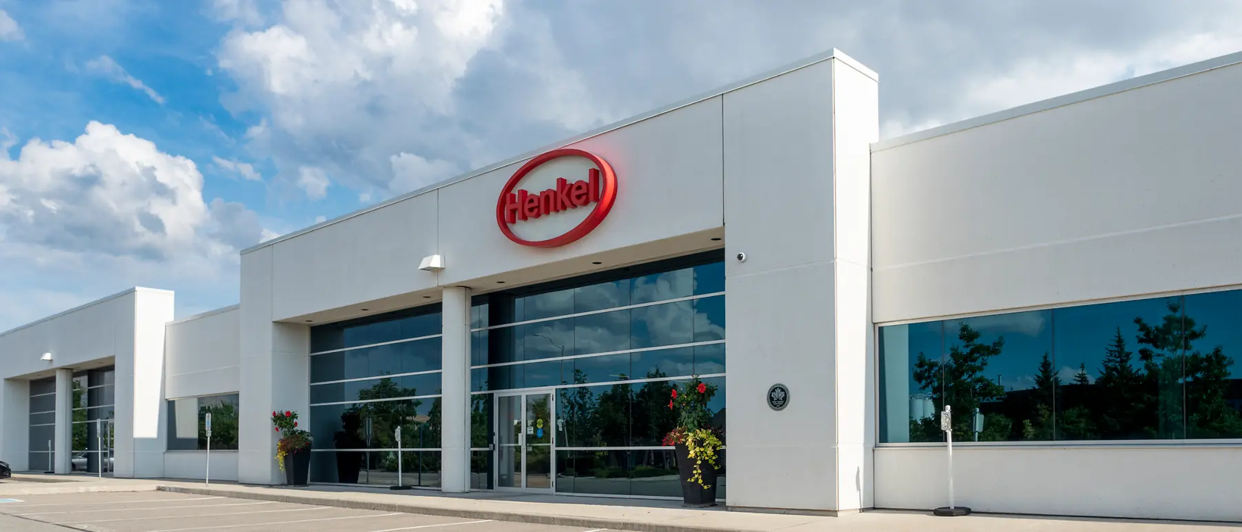 A modern Henkel North America office building with large glass windows, landscaped walkways, and the Henkel logo displayed above the main entrance.