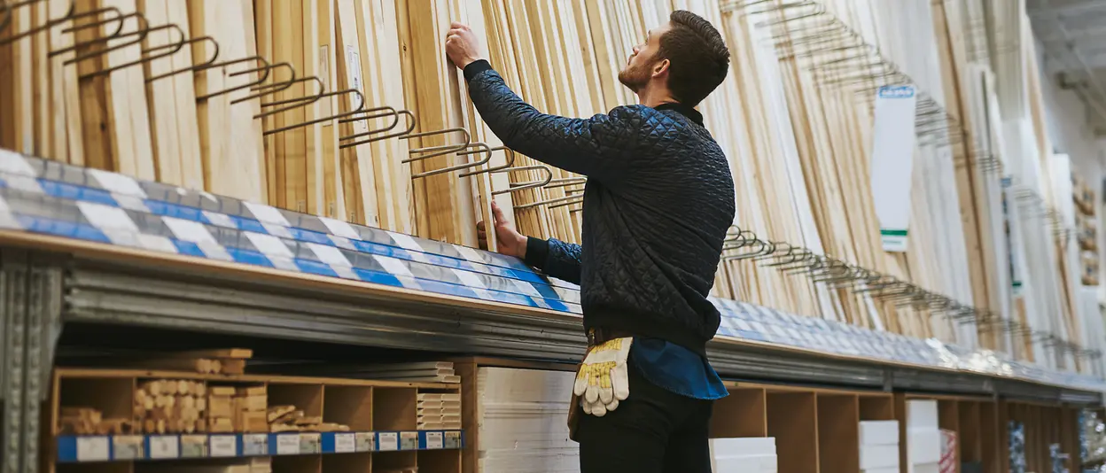 A man stands in front of a shelf of wooden slats in a building supply store.