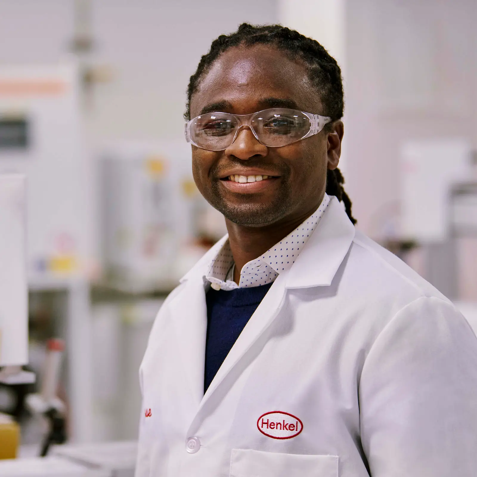A man in a lab coat with Henkel logo standing in a lab next to a microscope