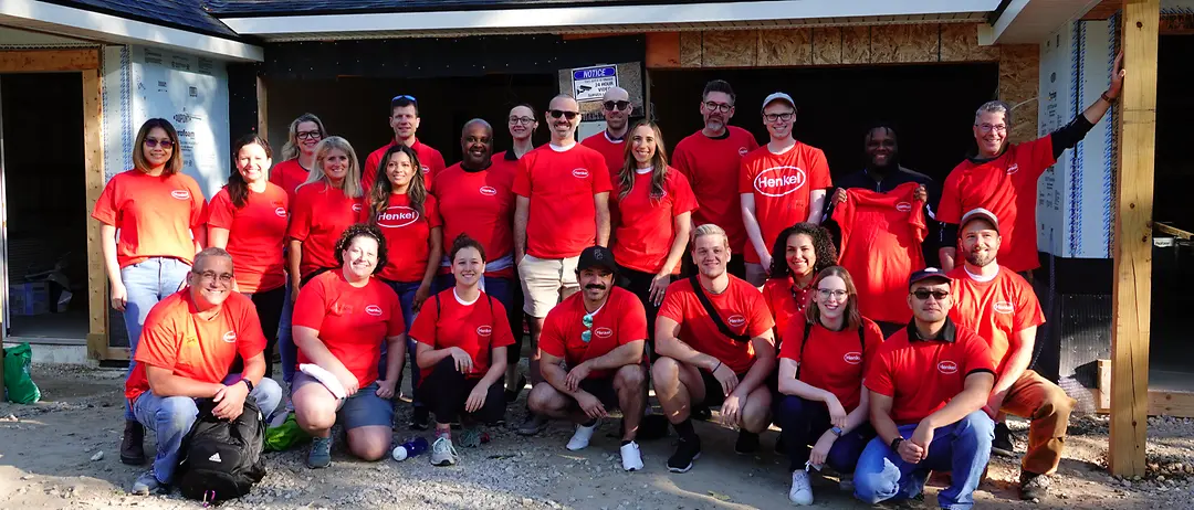 A group of Henkel employees and friends wearing red Henkel shirts standing together in front of the house they are building with Habitat for Humanity.