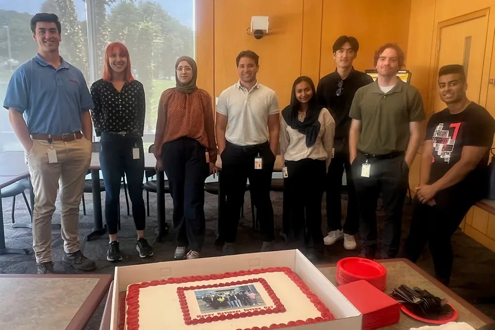 A small group of young people gather behind a cake decorated to celebrate National Intern Day