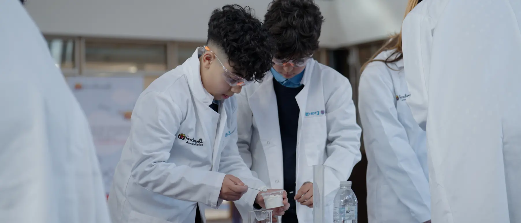 Two young boys wearing white lab coats and safety goggles conduct a science experiment.