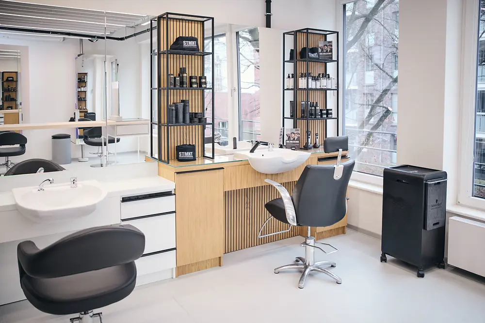 A bright, modern hair salon with two black styling chairs in front of shelves filled with hair care products and two white washbasins.