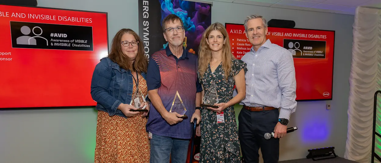 Bill and three colleagues standing together and holding awards with digital screens in background showing AVID logo.