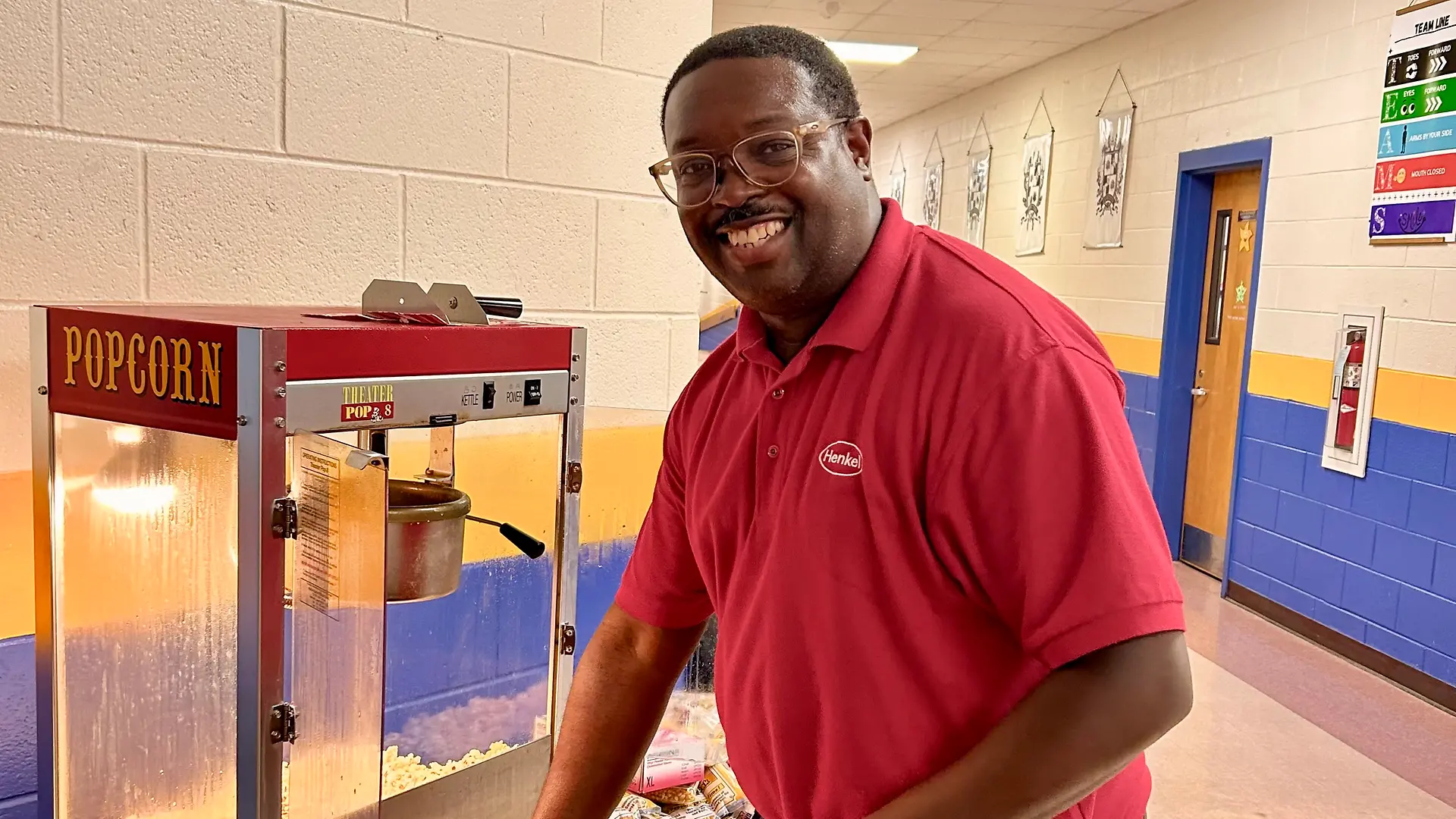 Man wearing a red photo shirt and gloves operating a popcorn machine in a blue hallway with yellow painted walls and tiled flooring.