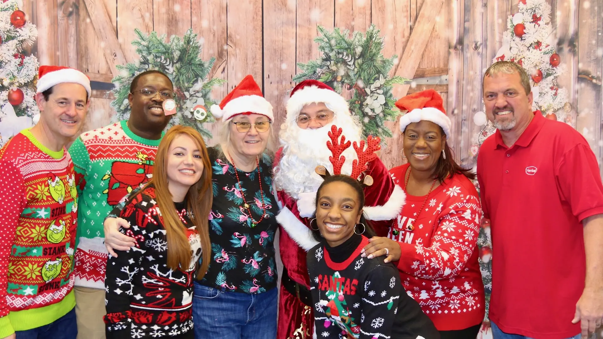 Group of employees posing with a person dressed as Santa Claus in front of a festive holiday backdrop featuring rustic wooden barn doors, and decorated holiday trees with white lights. Individuals are wearing festive holiday sweaters.