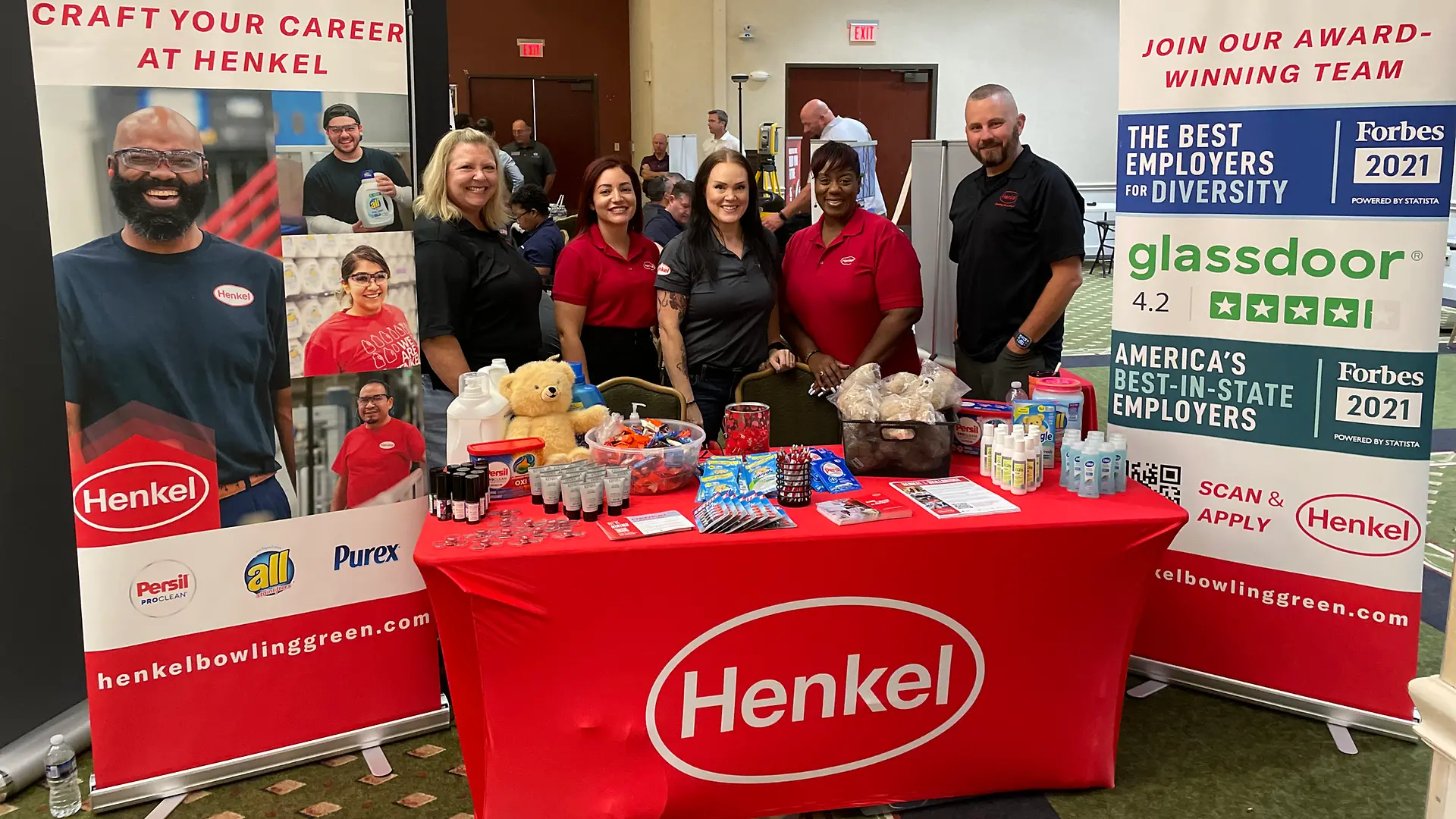 Team members stand behind a rectangular table covered with red Henkel branded table cloth. Behind the table are two vertical banners.