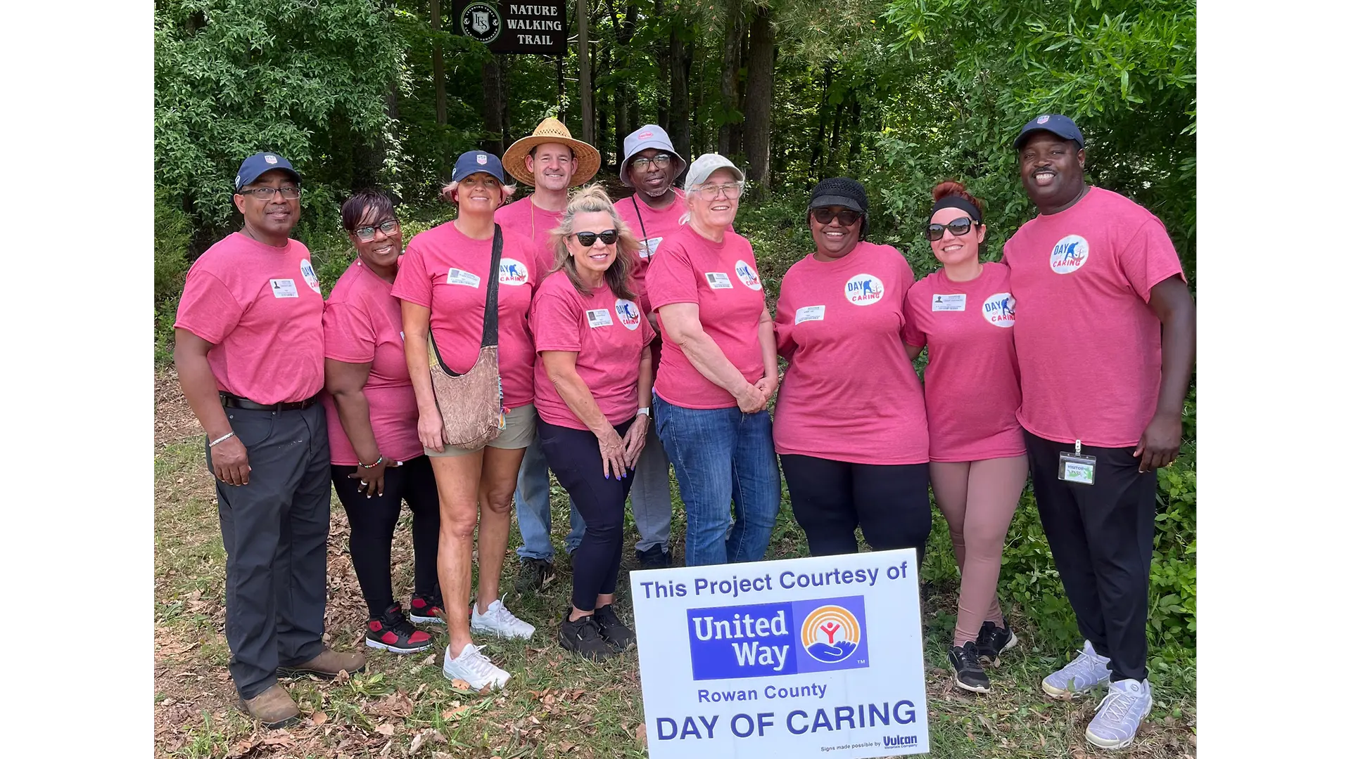Group of volunteers wearing matching pink t-shirts standing outdoors with a white sign that reads “This Project Courtesy of United Way.”