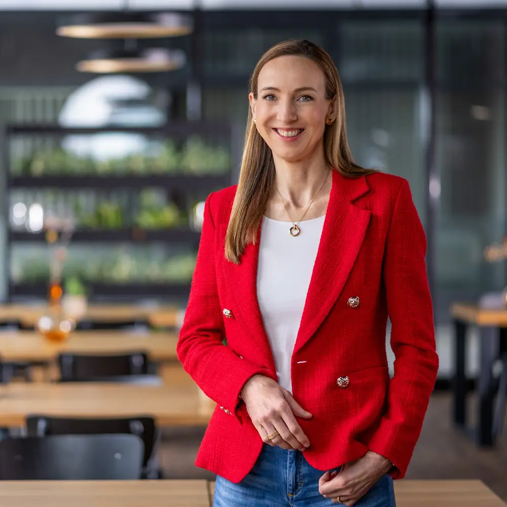 Simone Bagel-Trah博士身著亮紅色西裝外套和白色上衣，站在一個現代風格的咖啡館中，背景是木質桌子和裝飾花瓶。