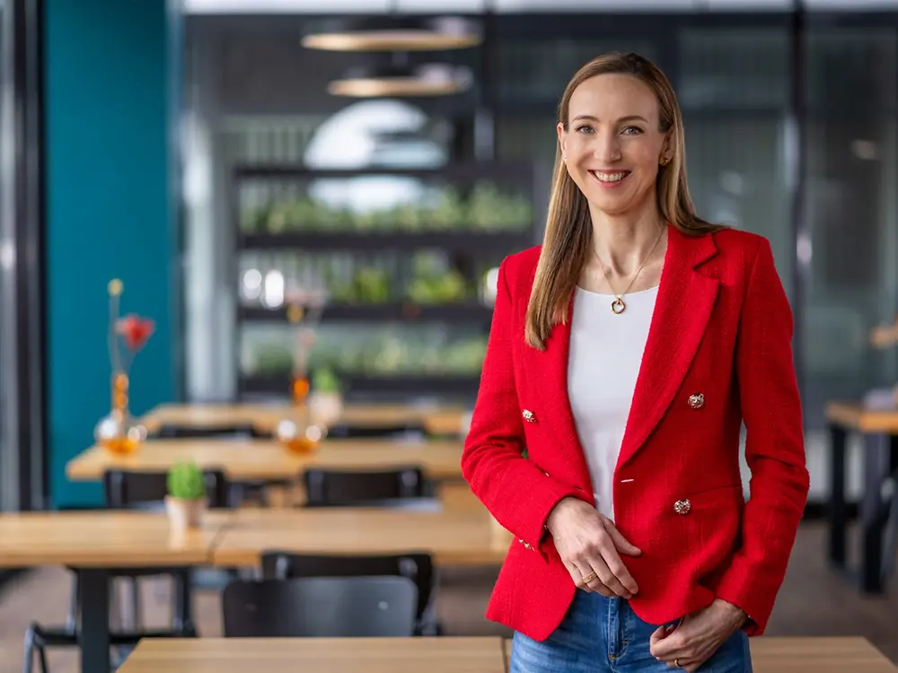 Dr. Simone Bagel-Trah wearing a bright red blazer and white top, standing in a modern café-style setting with wooden tables and decorative vases in the background.