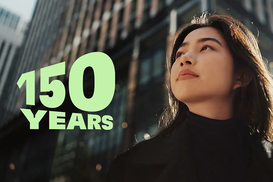 
Green text reading ‘150 YEARS’ over a modern cityscape with tall glass buildings and autumn trees, symbolizing the 150th anniversary celebration.
