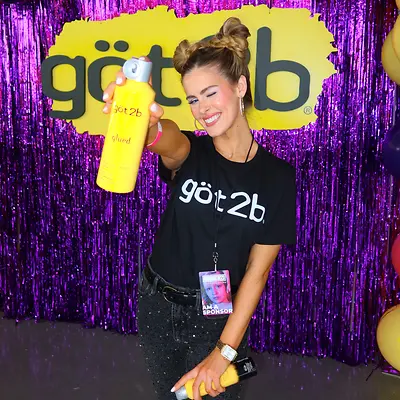 Woman holding got2b® Glued Blasting Freeze Spray, standing in front of sparkly purple background with got2b® yellow logo and balloons.