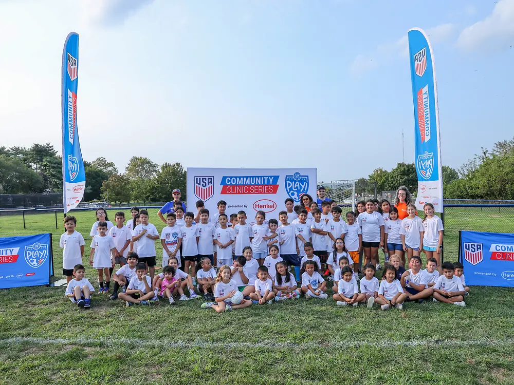 Large group of children and coaches posing on a soccer field at a U.S. Soccer community clinic, with banners behind them, during a youth event supported by Henkel to make soccer more accessible.
