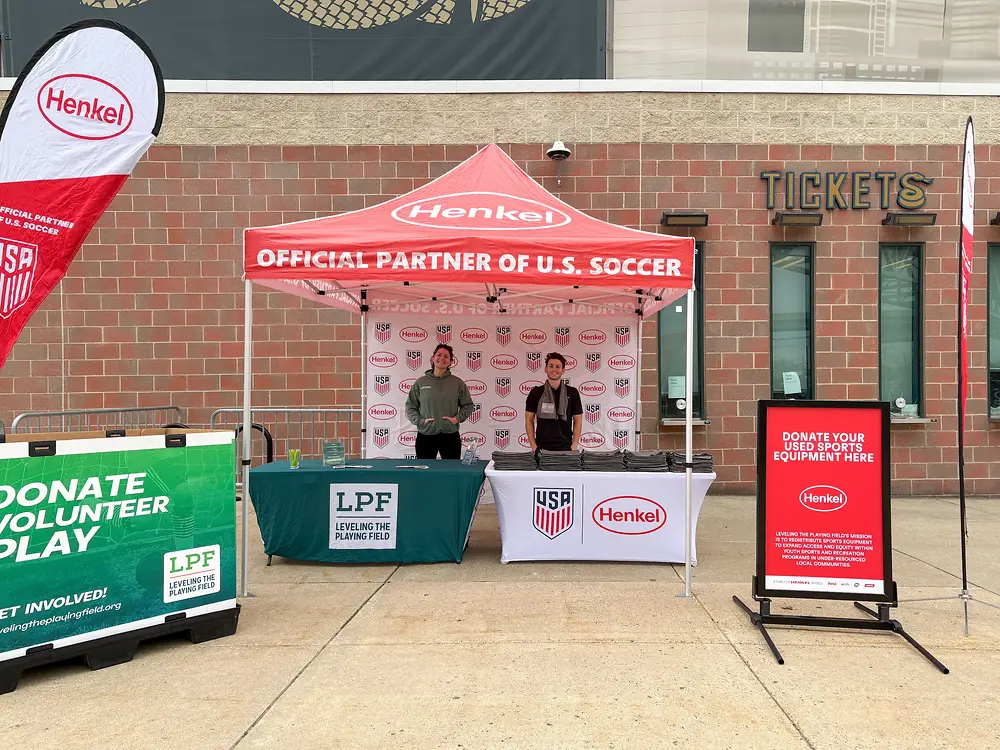 Outdoor event booth with a red Henkel canopy labeled 'Official Partner of U.S. Soccer,' two staff members standing behind tables with U.S. Soccer and Henkel logos, and promotional signs set up in front of a brick building.