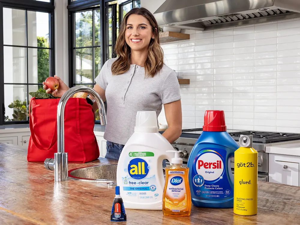 US soccer player Alex Morgan stands in a bright kitchen unpacking groceries from a red bag at the sink while several Henkel products including all free and clear detergent Persil detergent Dial soap got2b spray and a small glue bottle are displayed on the countertop.