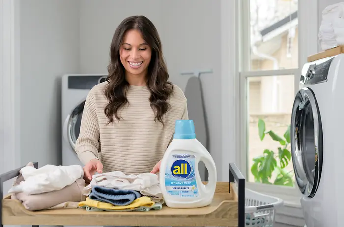 woman in laundry room folding clothes with all free clear bottle on the table