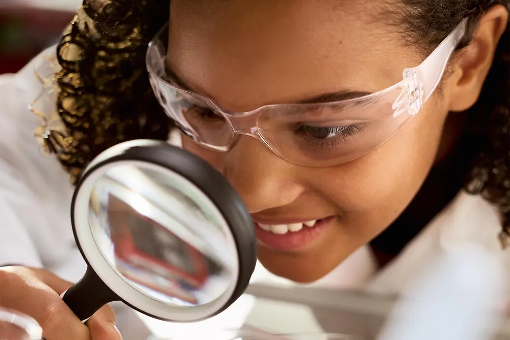 A young girl with safety goggles and a white lab coat looks at something through a magnifying glass.