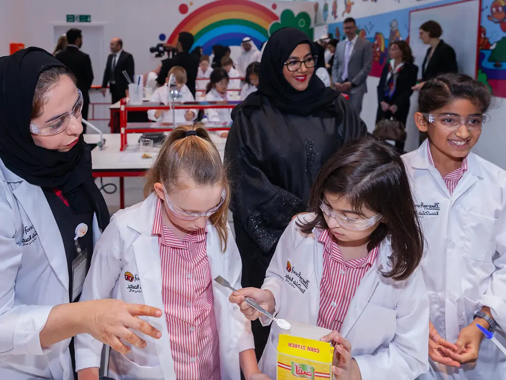 Several people in lab coats stand in a brightly decorated room next to a table where children are working with laboratory materials.