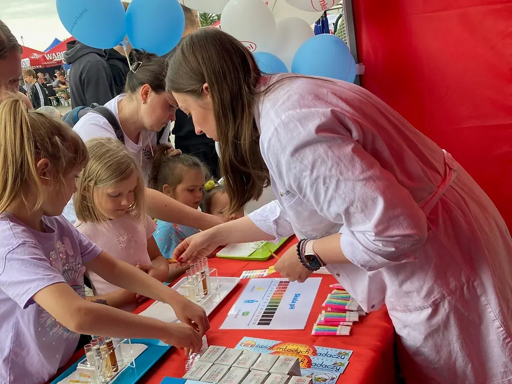 A person in a lab coat stands at a red booth and shows several children materials on a table.