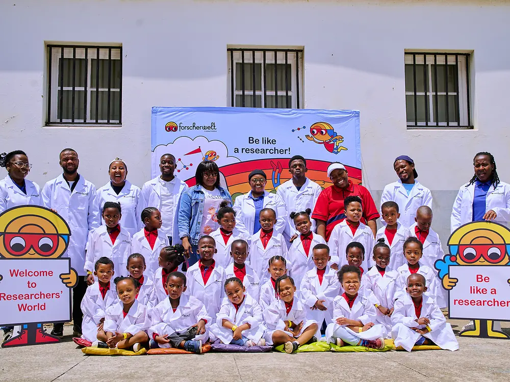 A group of adults and children wearing lab coats poses for a group photo. In front of them are two stands with Researchers’ World illustrations.