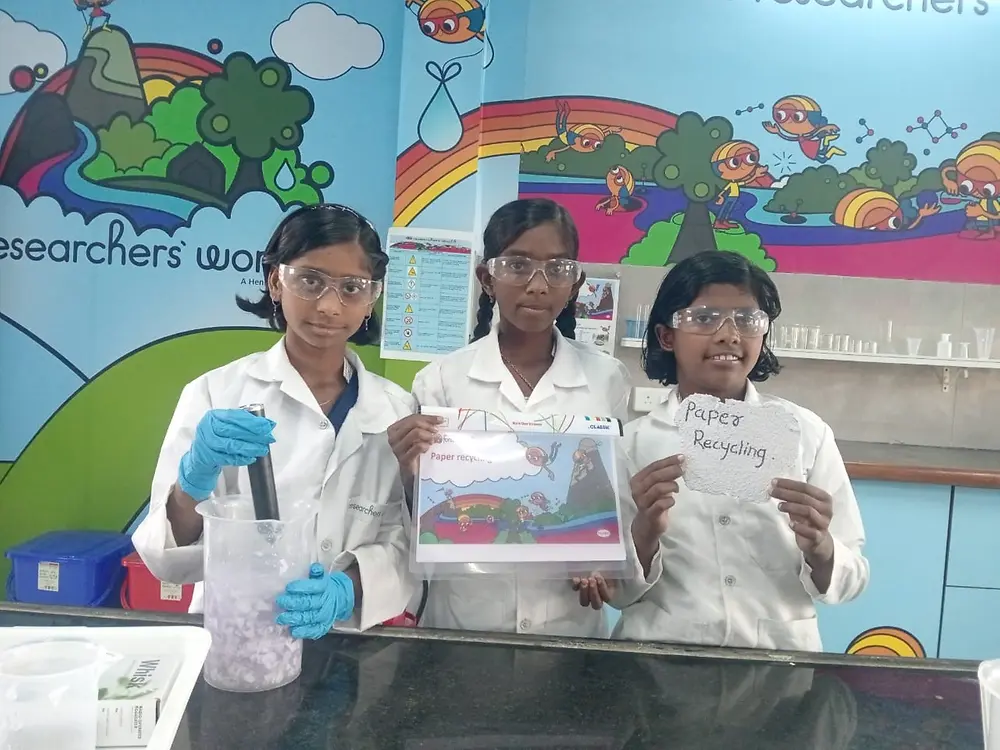 Three children in lab coats stand in a brightly decorated room. One person is holding a container, one is showing a poster, and one is holding a sheet of paper labeled ‘Paper Recycling.’