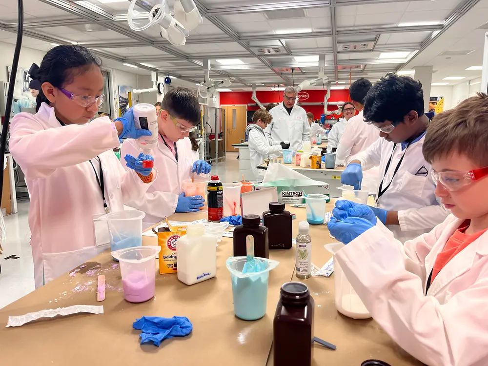 Several children in lab coats are working at a large table in a laboratory. Various containers, measuring cups, and colorful liquids are placed on the table.