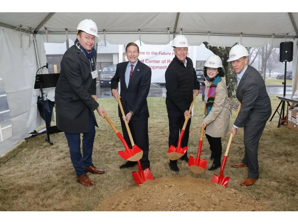 Group wearing hard hats break ground with red shovels under a tent at a Henkel facility event.