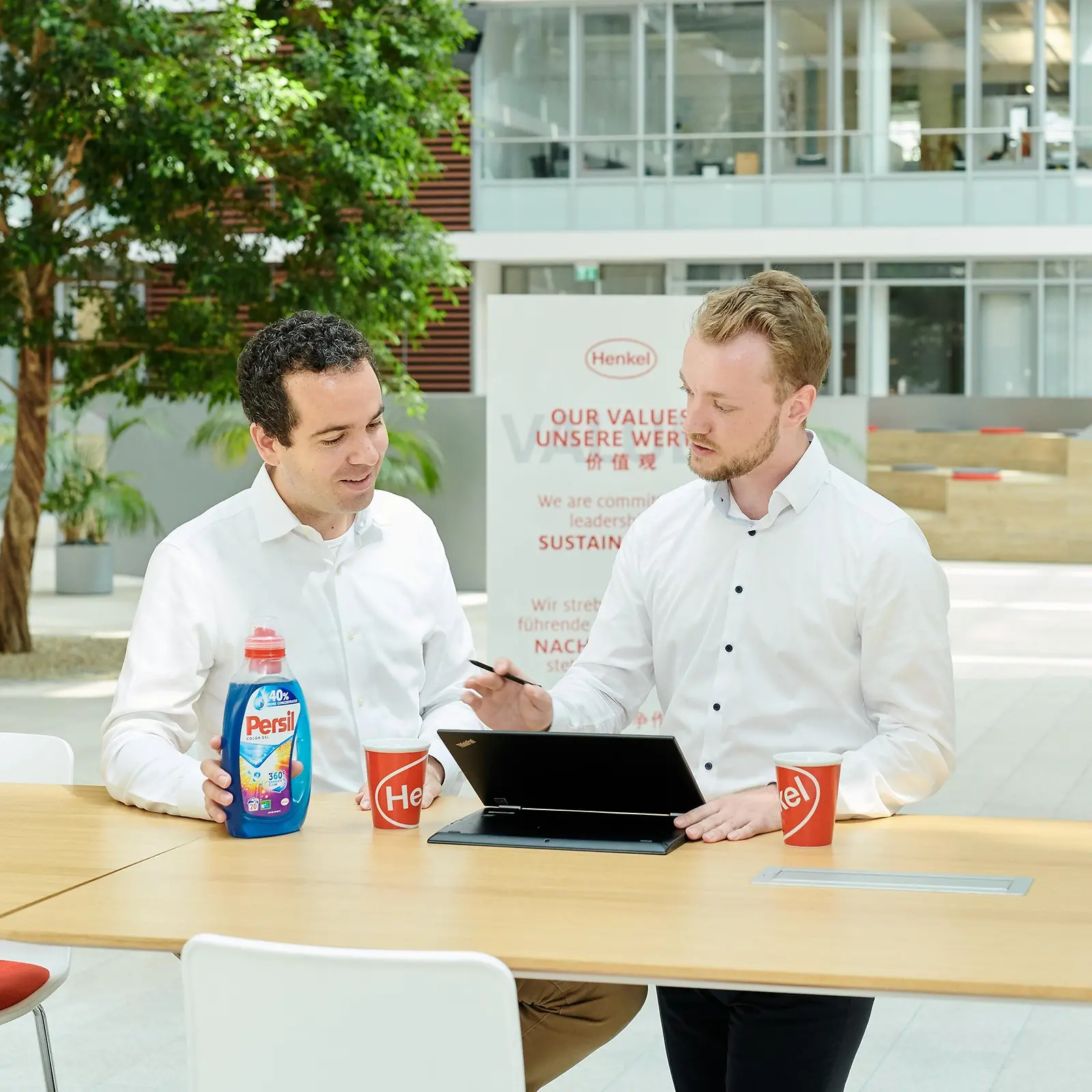 Two men standing at a table with a Persil bottle and a laptop