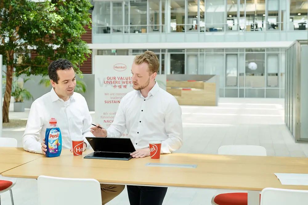 Two men standing at a table with a Persil bottle and a laptop